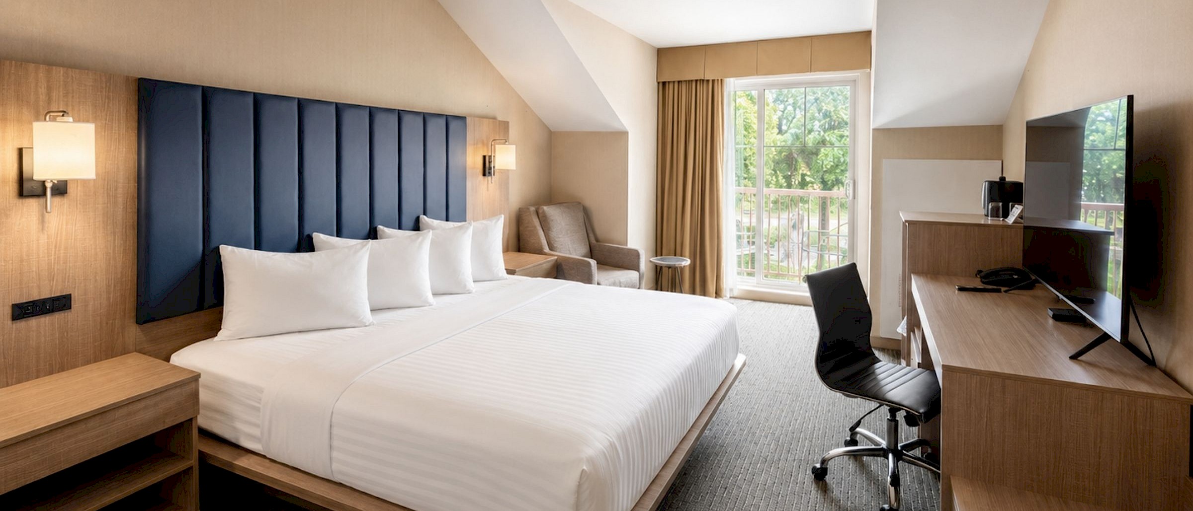 Spacious hotel king room with a navy blue headboard, work desk, and a private balcony overlooking lush greenery.