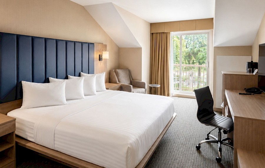 Spacious hotel king room with a navy blue headboard, work desk, and a private balcony overlooking lush greenery.