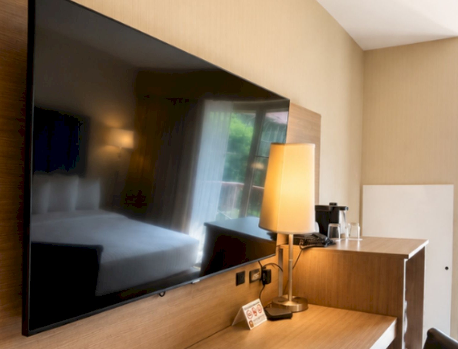 Large flat-screen TV mounted above a wooden desk setup in a modern hotel room with a balcony view.