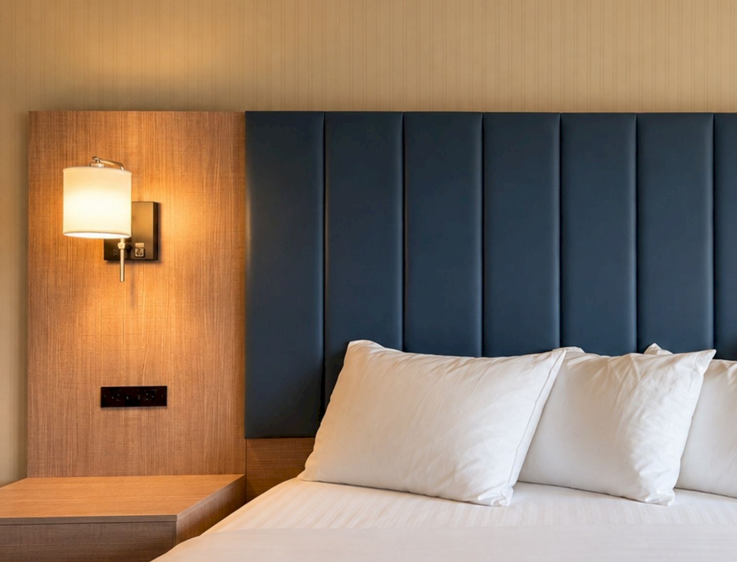 Close-up of a hotel king bed with white pillows, a navy blue padded headboard, and wood side paneling with wall sconces.